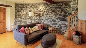 A cozy living room with a stone wall, a wooden staircase, and a grey sofa with multiple cushions. - Squirrel Cottage (Keswick)