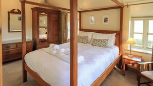 A four-poster bed with white linens and towels, a wooden wardrobe, a bedside table with a lamp, and a window with curtains. - Squirrel Cottage (Keswick)