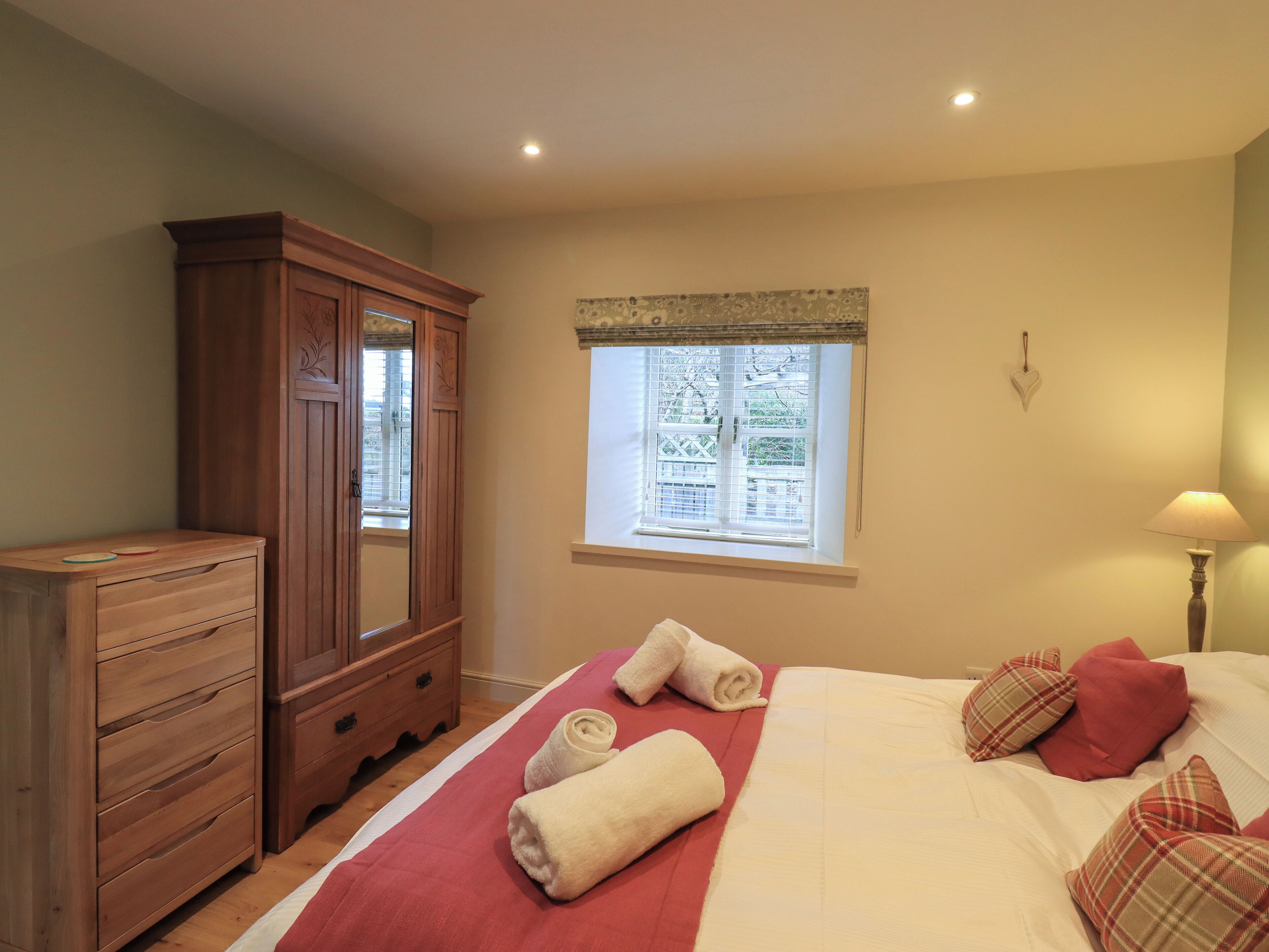 A bedroom with a wooden wardrobe, a window with blinds, a bed with red and white bedding, and a bedside lamp.
