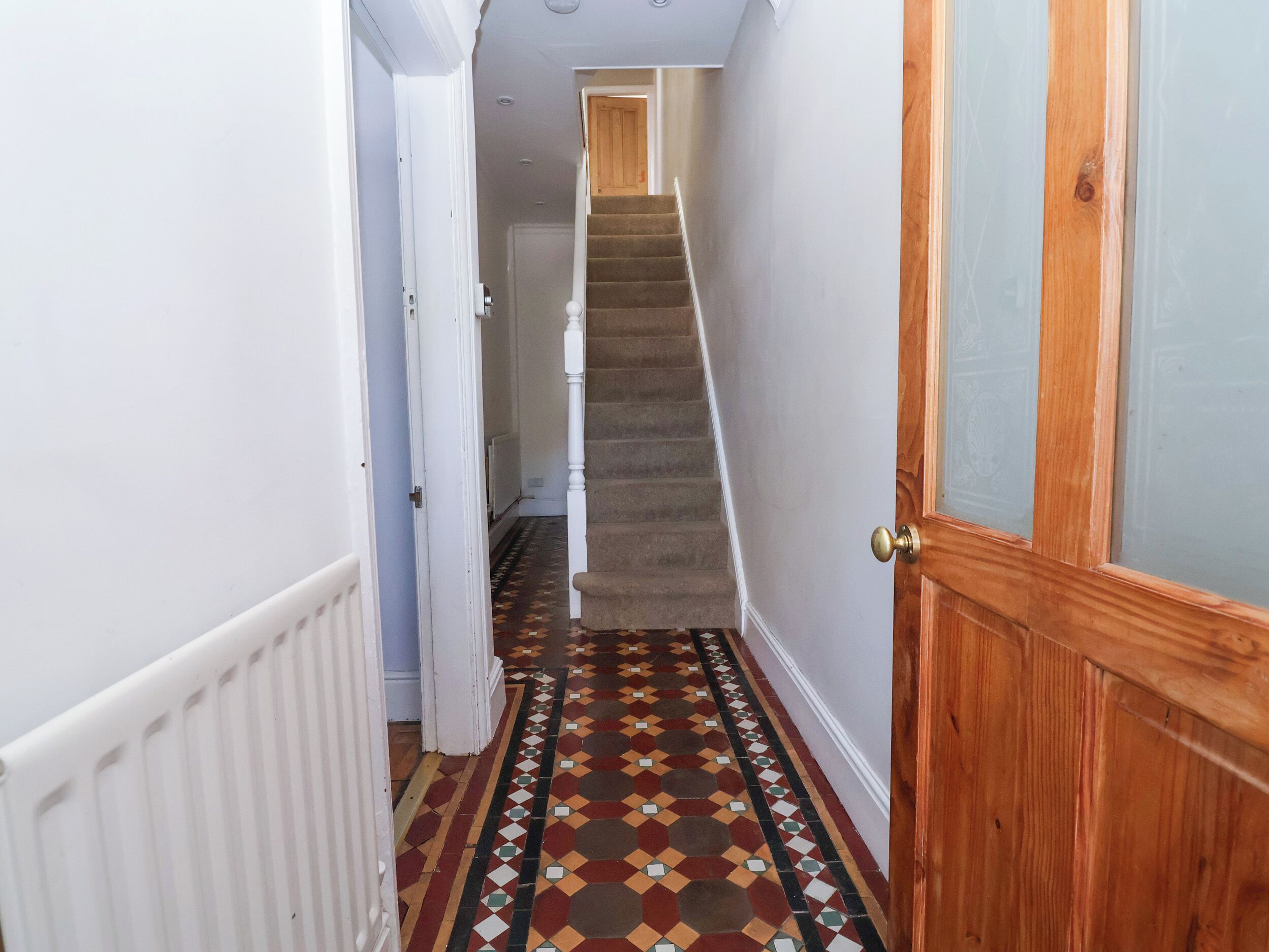A narrow hallway with a patterned floor, a wooden door, and a staircase leading to another room.
