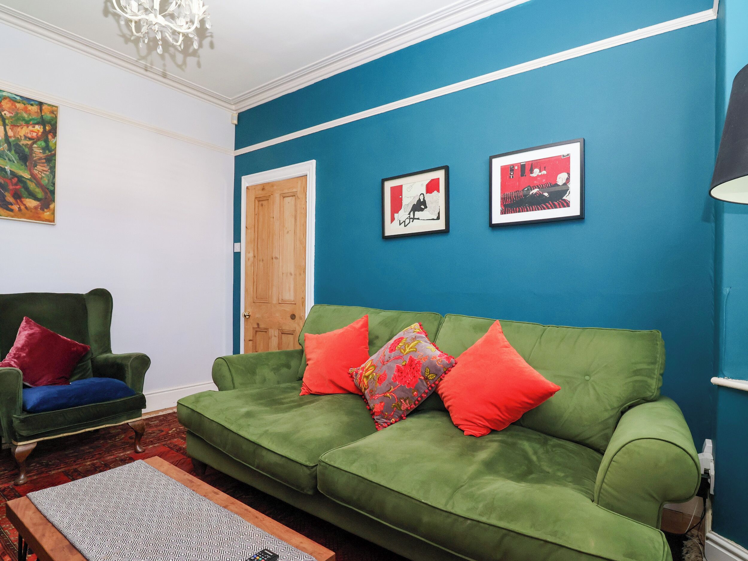 Een woonkamer met een groene bank, rode en kleurrijke kussens, een houten deur en twee ingelijste kunstwerken aan de muur.