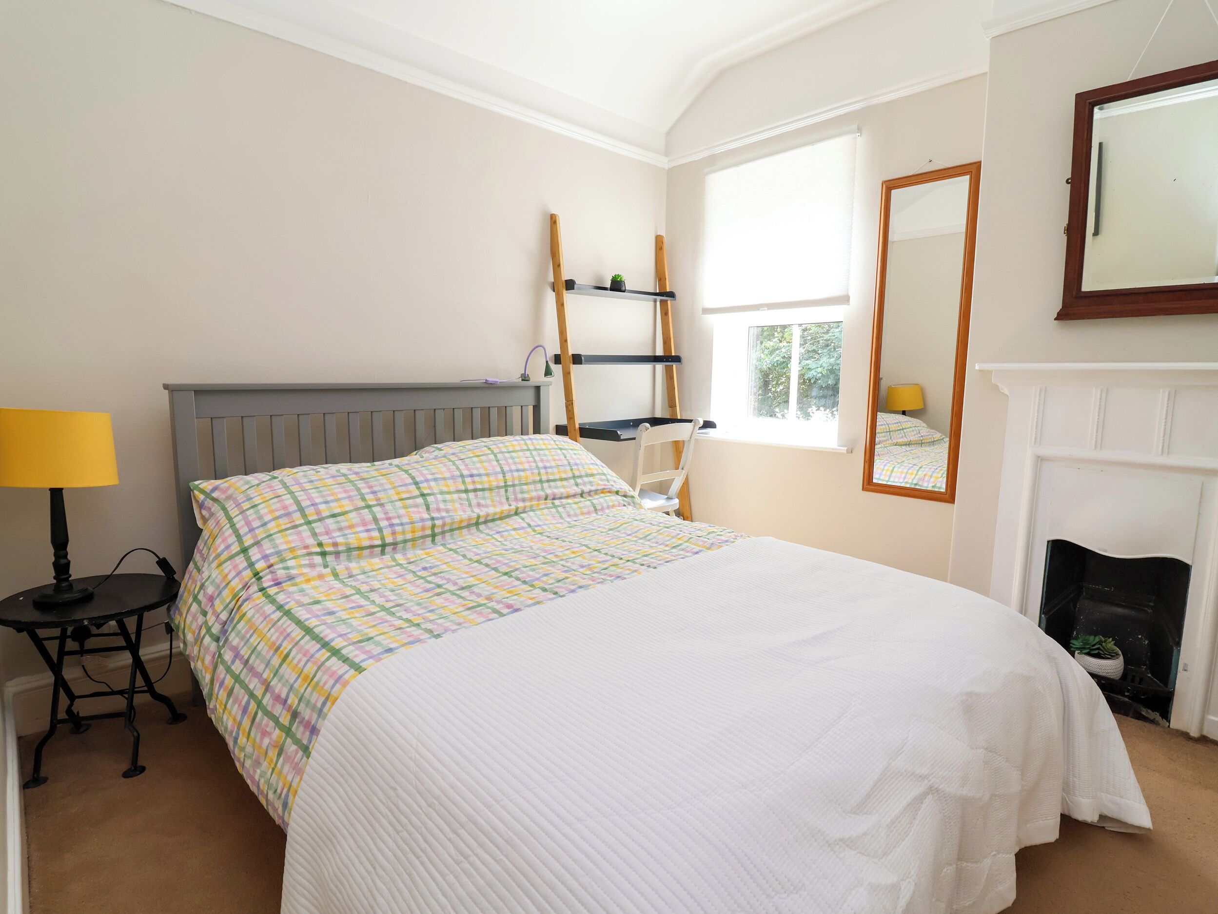 A bedroom with a bed, a desk, a chair, a window, a mirror, and a fireplace.
