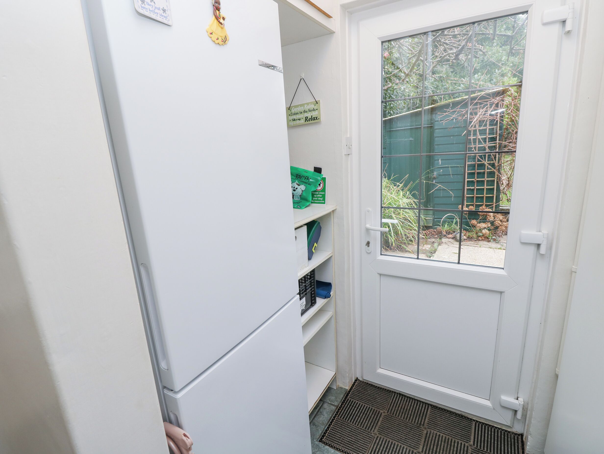 A compact kitchen with a white refrigerator, a door with a window, and a small shelf with green boxes.