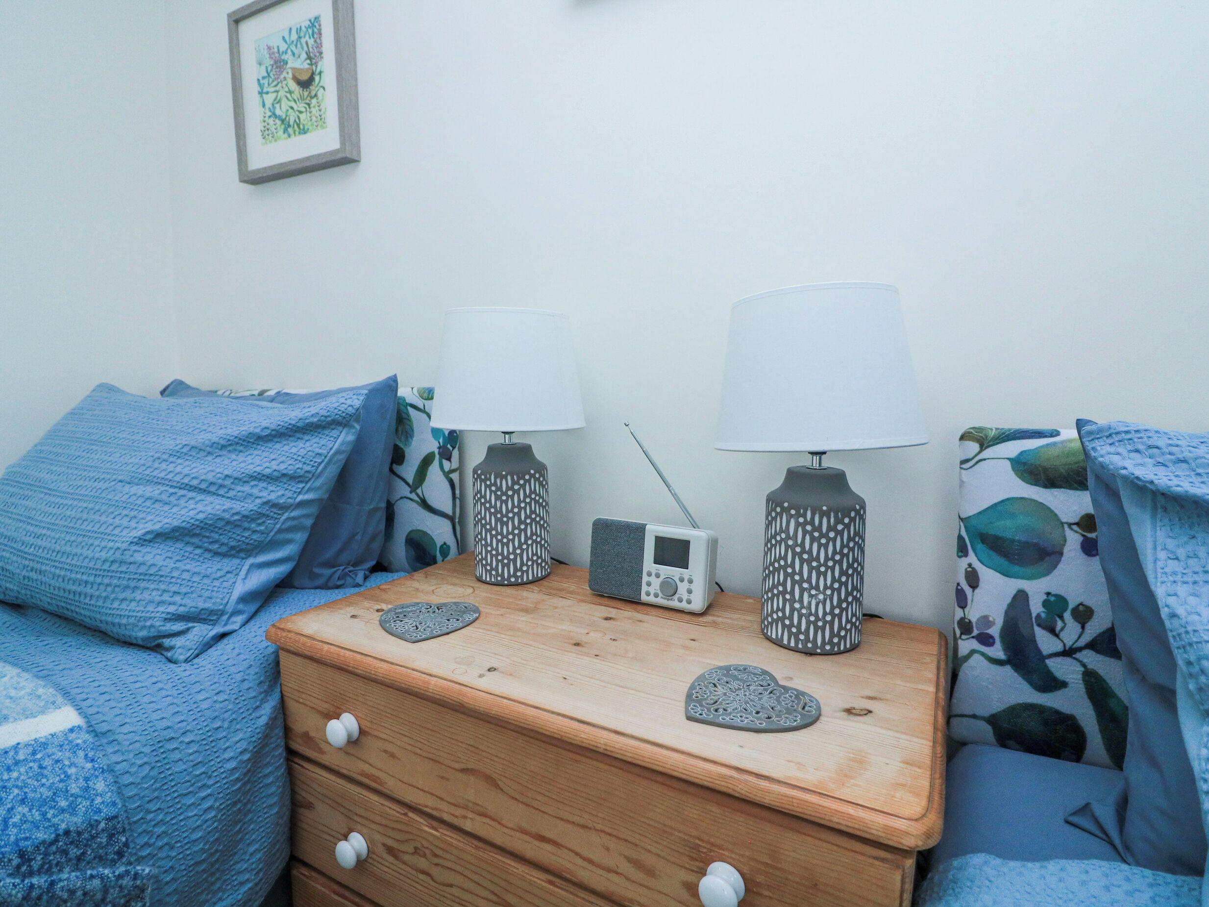 A neatly made bed with blue bedding, a wooden nightstand with two lamps, a radio, and decorative items.