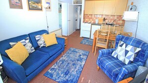 Un espacio habitable compacto con un sofá azul, un sillón escocés, una zona de cocina y una mesa de comedor.