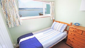 Una cama individual con una colcha a rayas, un aparador de madera con cajones, una ventana con cortinas y vistas a un cuerpo de agua y tierra.