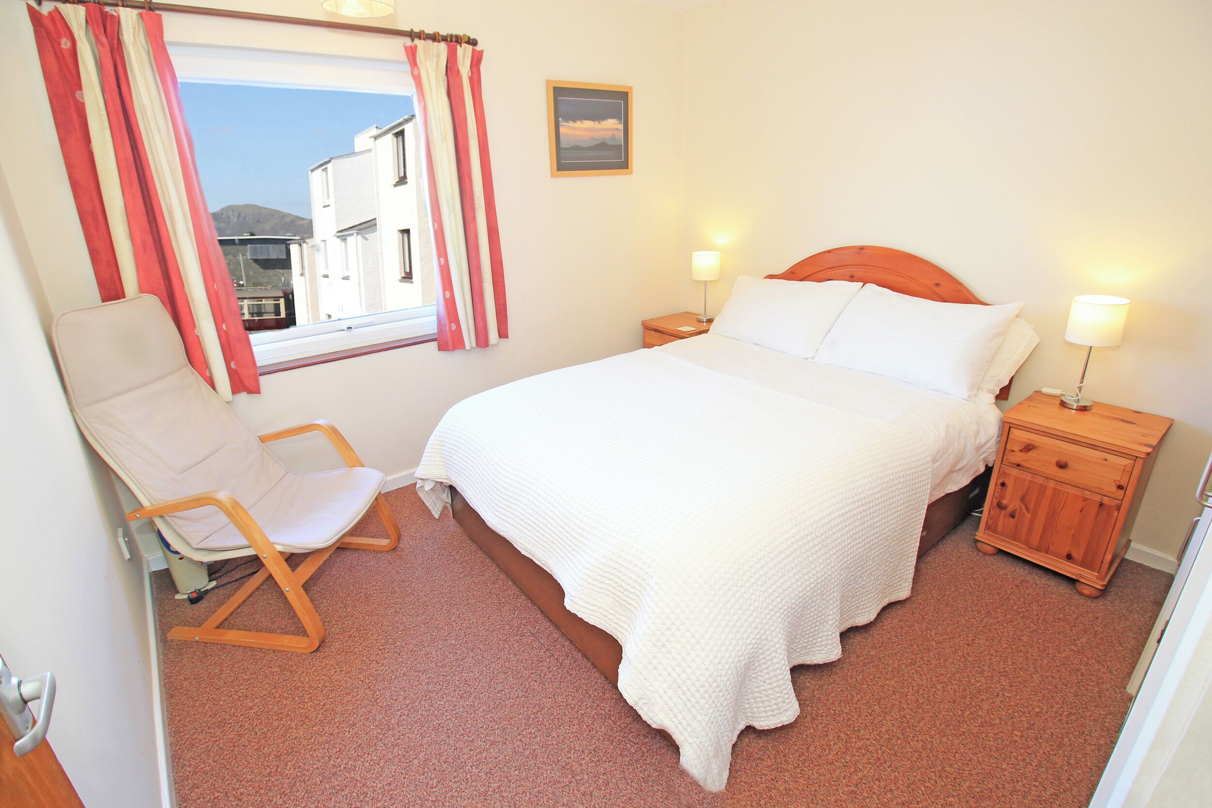 A hotel room with a bed, a chair, a nightstand, a lamp, a window with curtains, and a picture on the wall.