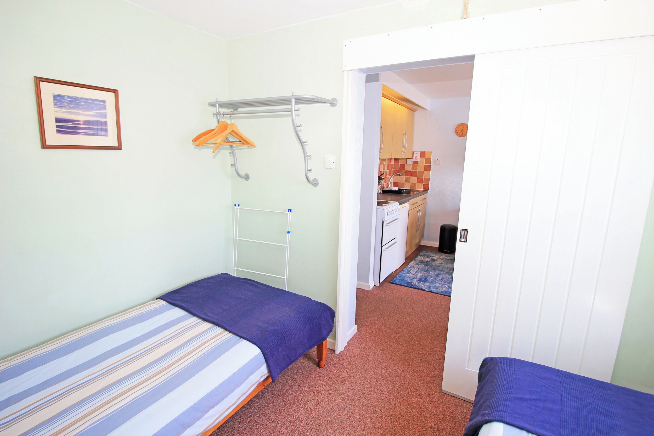 A small, well-lit room with two single beds, a hanging clothes rack, and a view into a kitchen area.