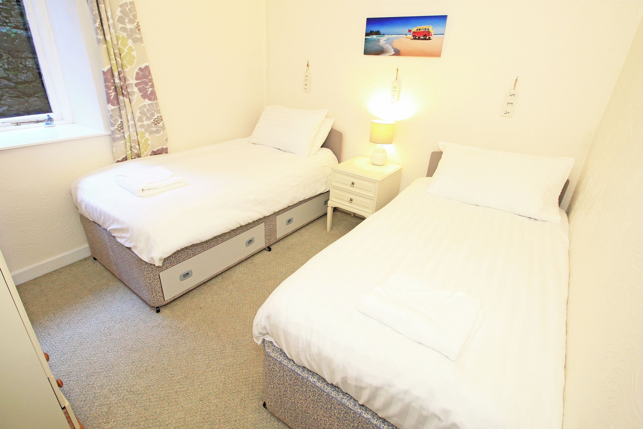 A twin room with two beds, a nightstand with a lamp, a window with curtains, and a picture on the wall.