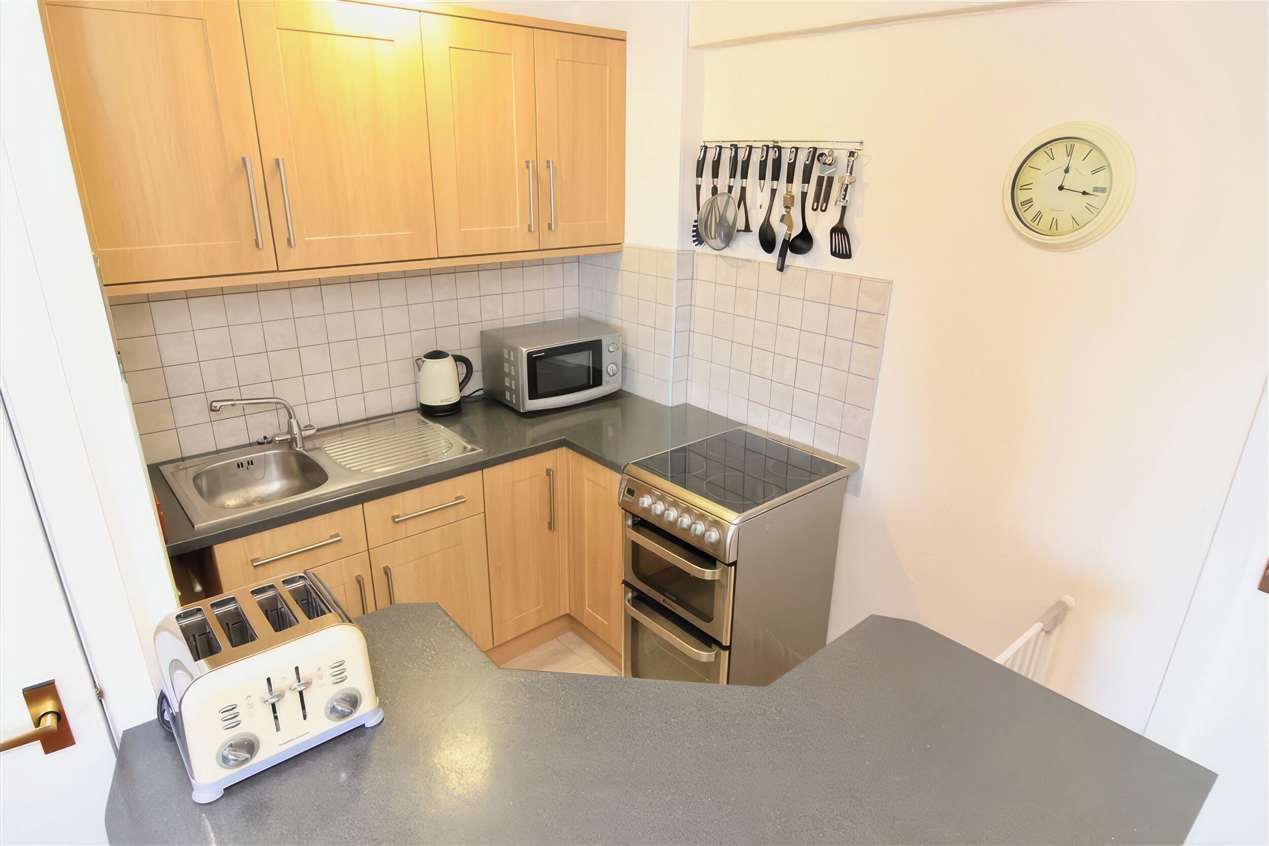 A compact kitchen with wooden cabinets, a microwave, a toaster, and a clock on the wall.