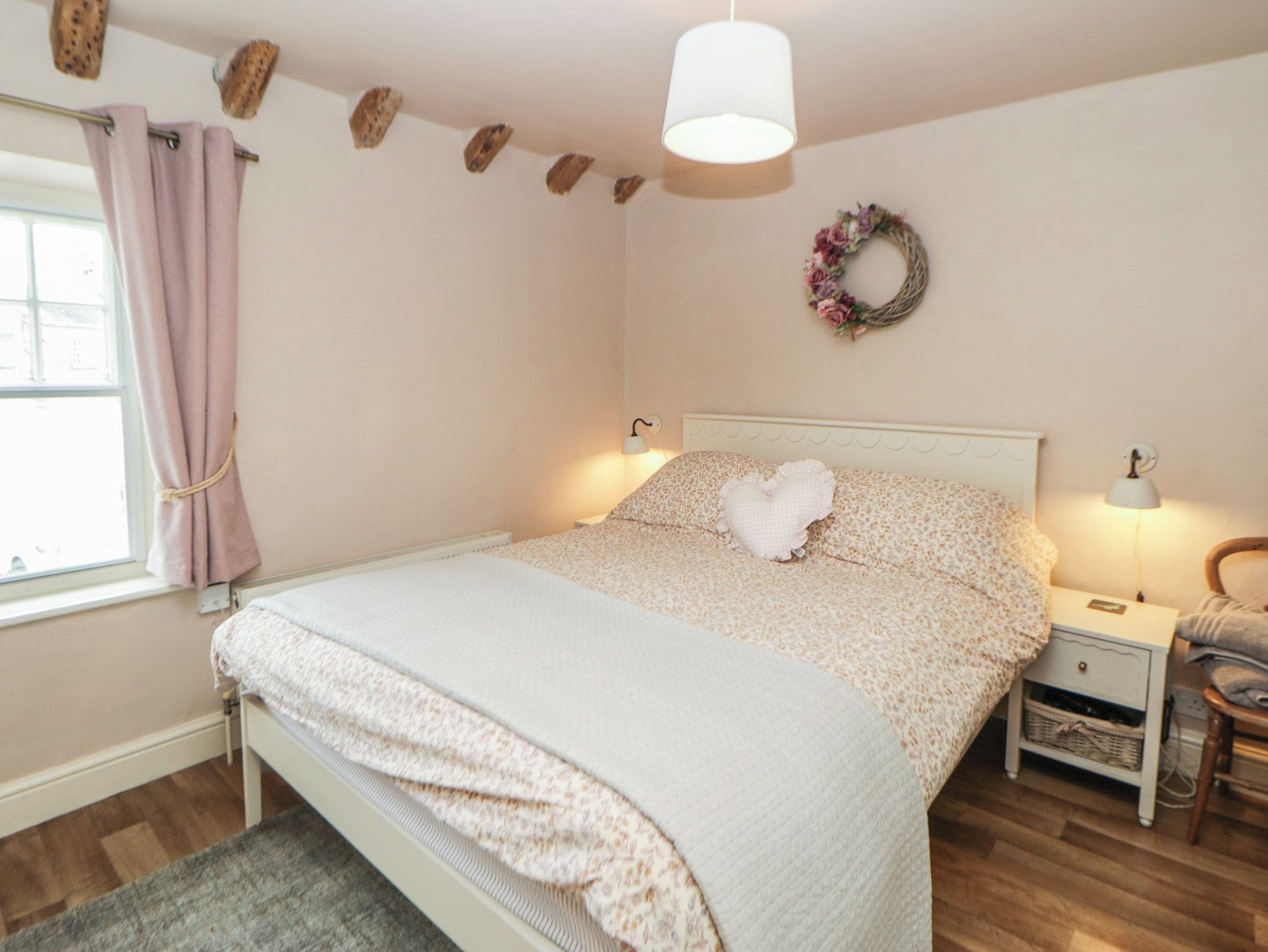A bedroom with a bed, bedside tables, a nightstand, a window with curtains, a wall-mounted lamp, and a wreath.
