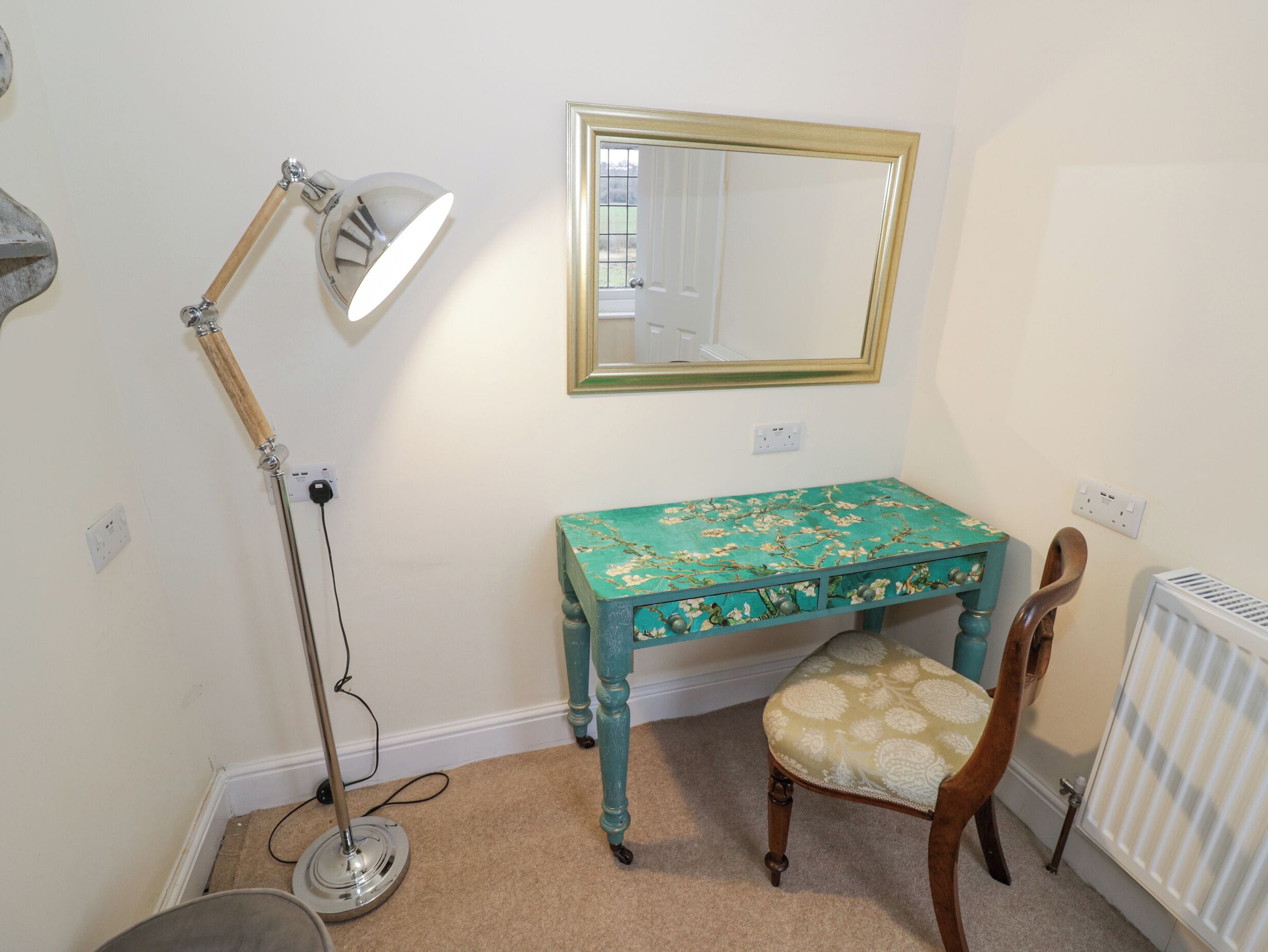 A room with a teal vanity table, a chair, a floor lamp, a mirror, and a radiator.