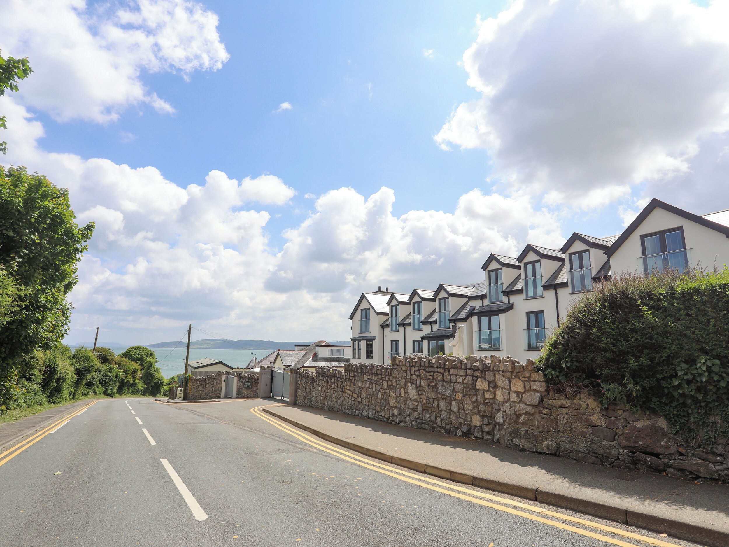 A coastal road with houses, a stone wall, and a clear sky.