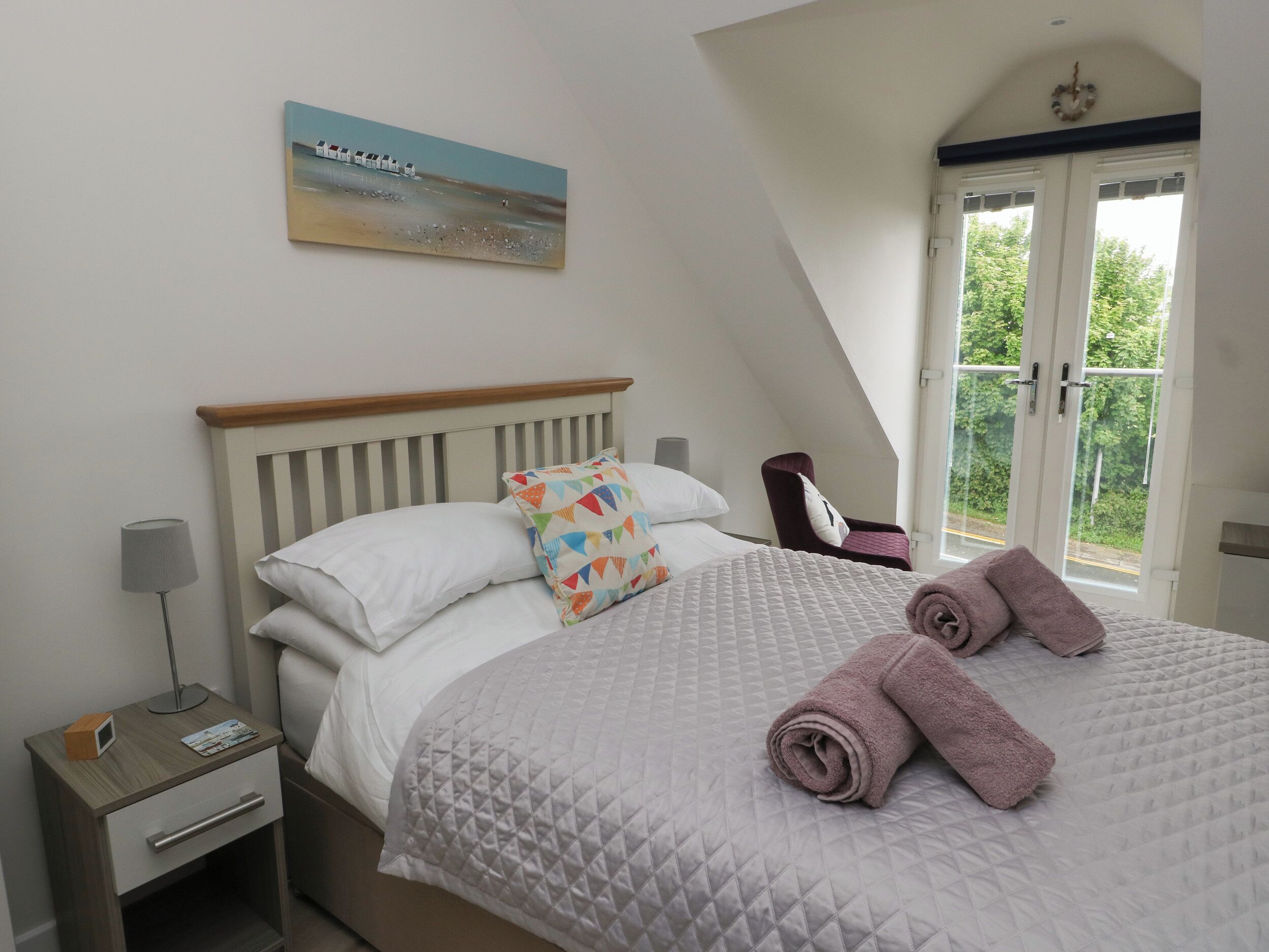 A bedroom with a bed, a nightstand, a lamp, a chair, and a window with a view of greenery.