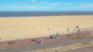 Een breed zandstrand met een helderblauwe lucht, enkele mensen die wandelen, en een omheind pad met bankjes.