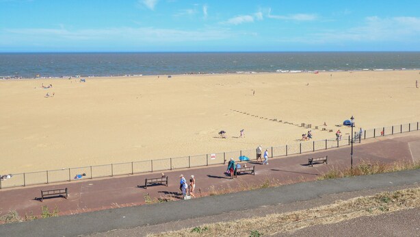 A wide sandy beach with a clear blue sky, a few people walking, and a fenced walkway with benches. - Tiffany's Cottage (Great Yarmouth)