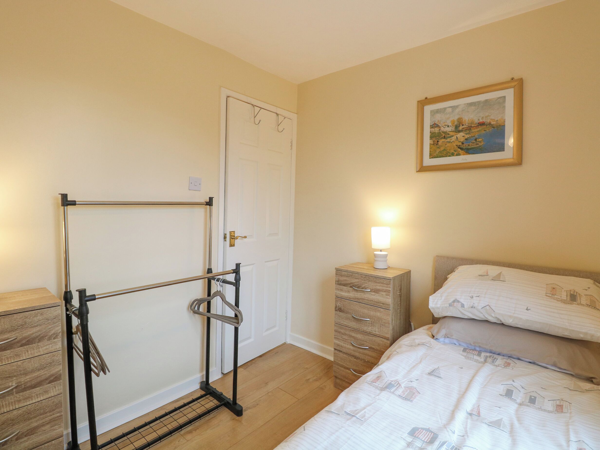 A bedroom with a bed, a nightstand with a lamp, a wooden dresser, a door, and a wall-mounted rack.
