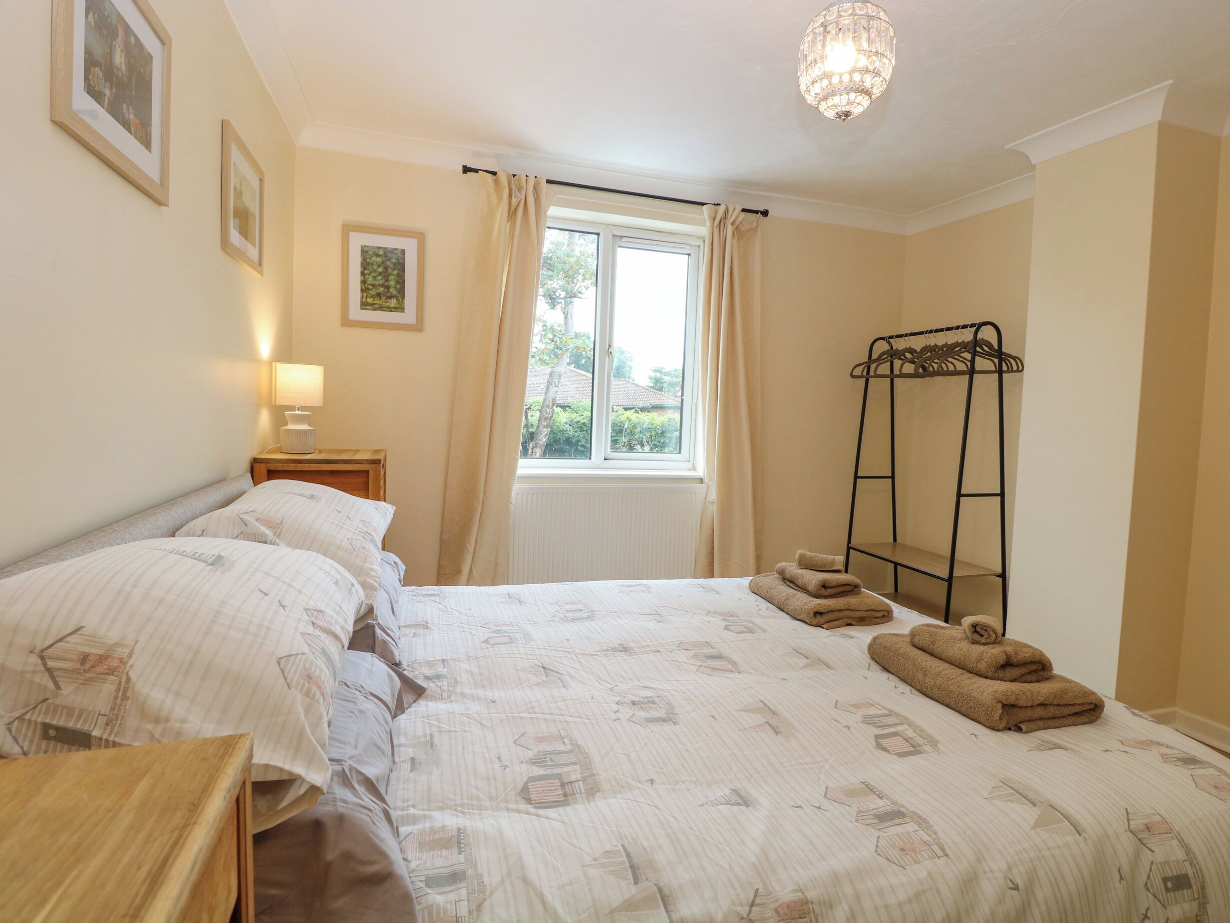 A bedroom with a bed, a bedside table with a lamp, a window with curtains, and a metal drying rack.