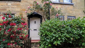 A stone building with a white door, surrounded by lush greenery and flowering plants. - Rokeby House (Whitby)