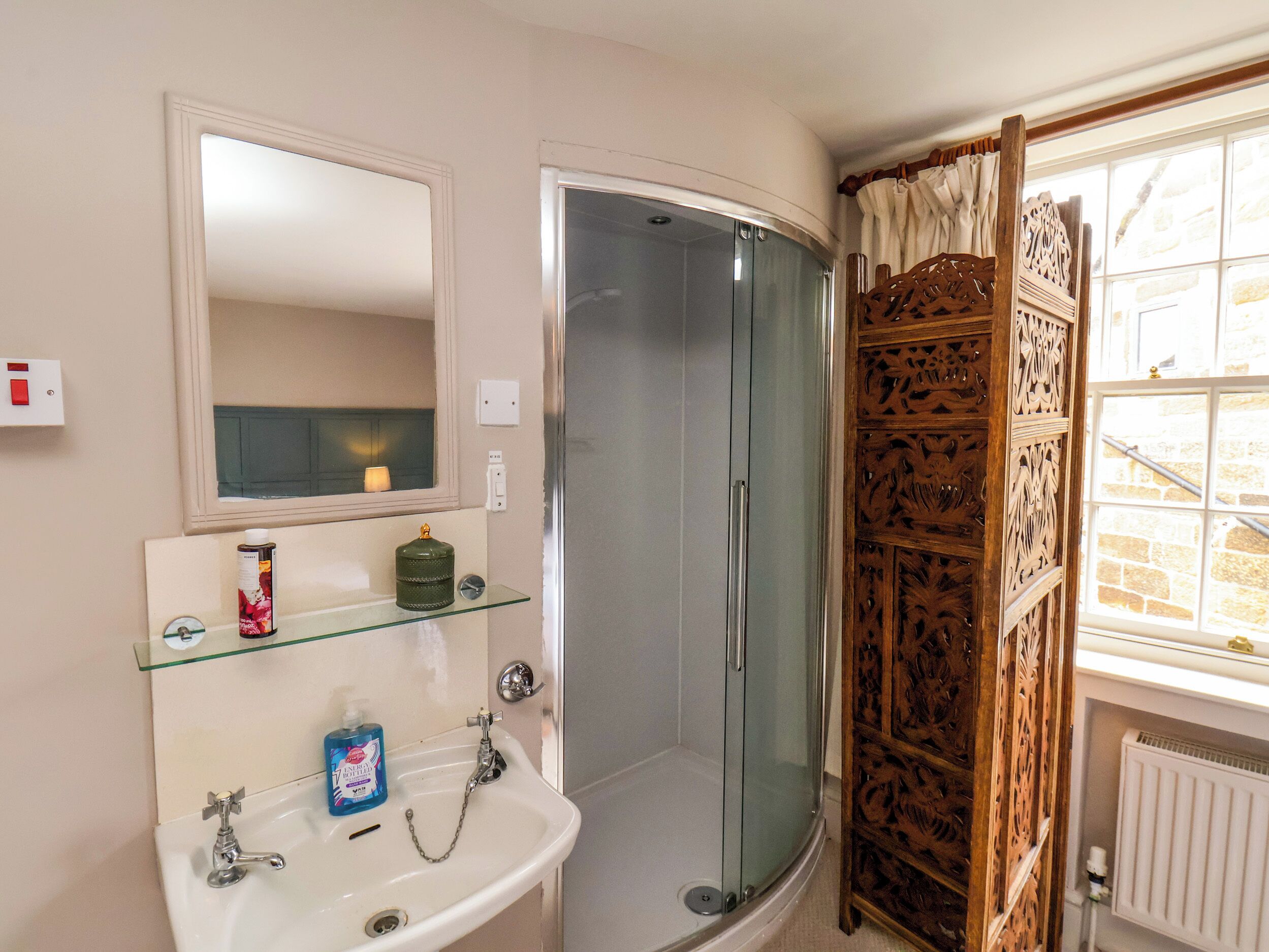 Une salle de bain avec un lavabo blanc, une cabine de douche en verre, un miroir et un panneau décoratif en bois près de la fenêtre.