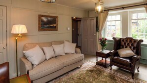 A living room with a sofa, armchair, and a lamp. There is a painting on the wall and a door leading to another room. - Rokeby House (Whitby)