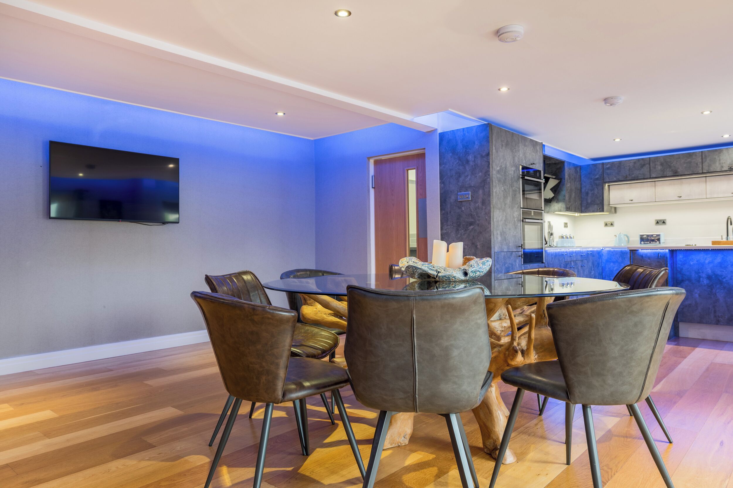 Un espace repas moderne avec une table ronde et des chaises en cuir, un téléviseur à écran plat fixé au mur et une cuisine dotée d’un éclairage bleu.