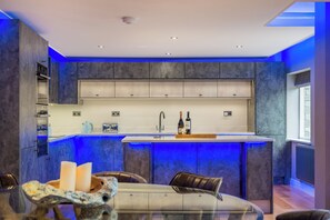A modern kitchen with blue lighting, a central island, and a dining area with a glass table and chairs. - Ullswater View House (Penrith)