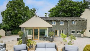 A patio with two grey sofas and a wooden table, a stone building with a sloped roof, and a garden with various plants. - Ullswater View House (Penrith)