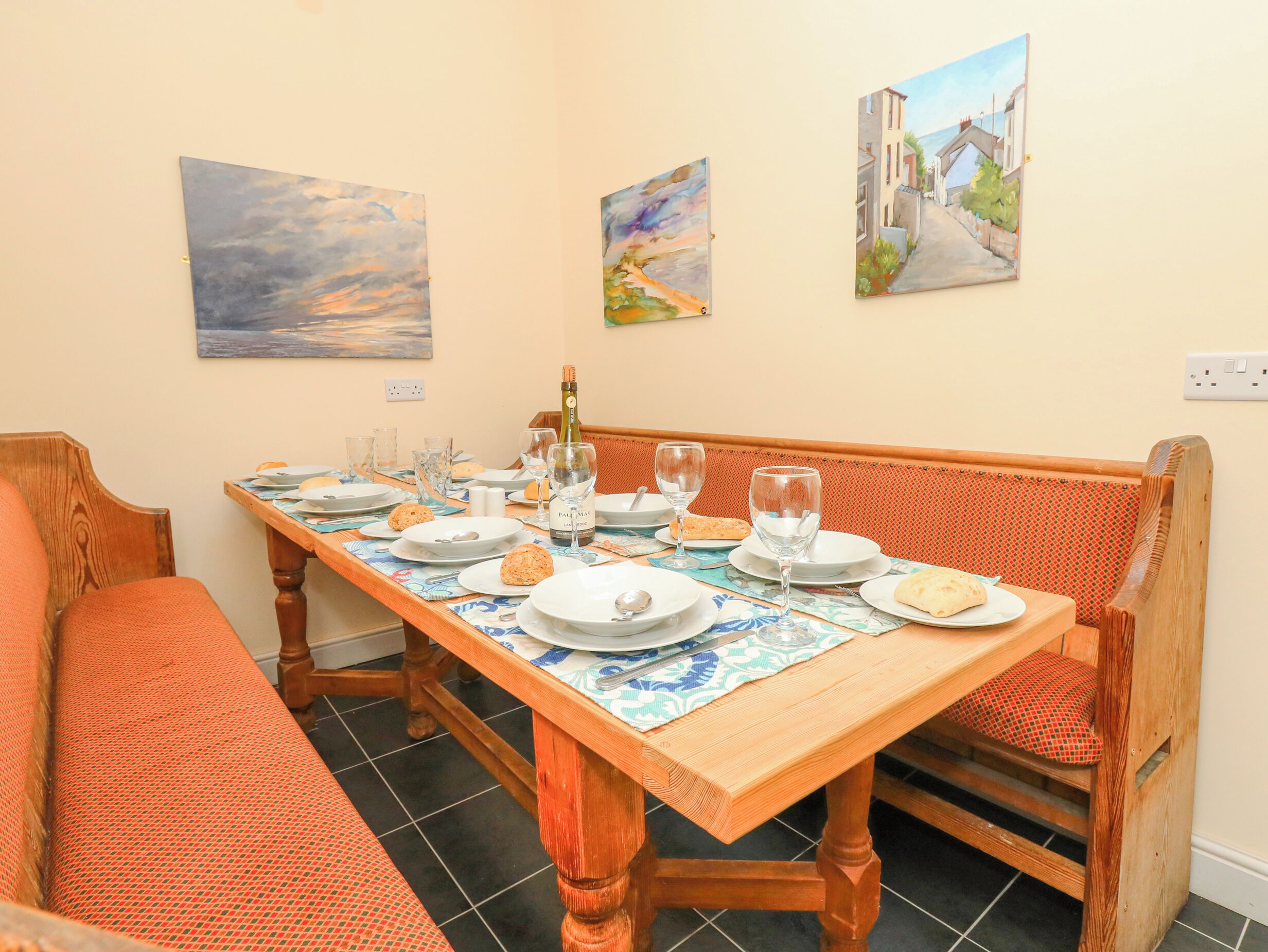 A dining area with a wooden table set for four, surrounded by orange cushioned benches. There are three paintings on the wall above the table.
