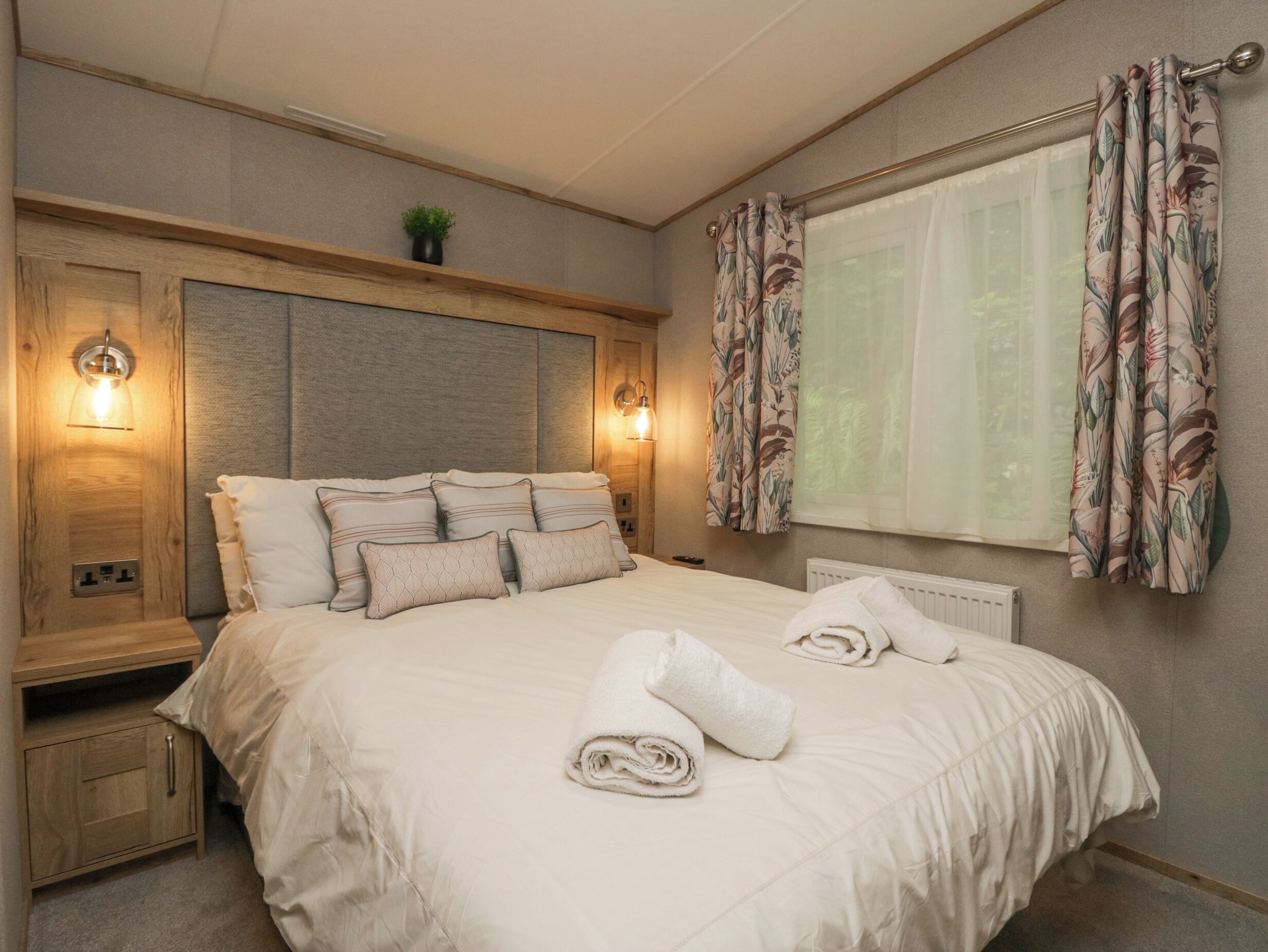A neatly made bed with a headboard, bedside lamps, a nightstand, and a window with curtains.