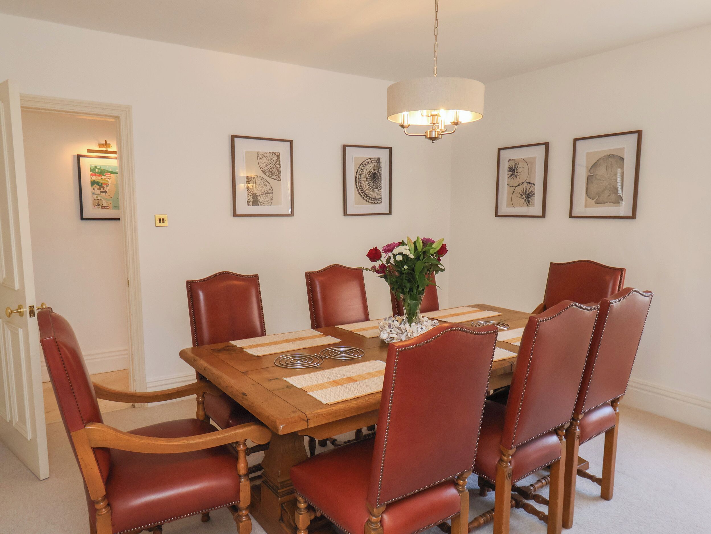 A dining room with a wooden table, six red leather chairs, a vase with flowers, and framed artwork on the walls.