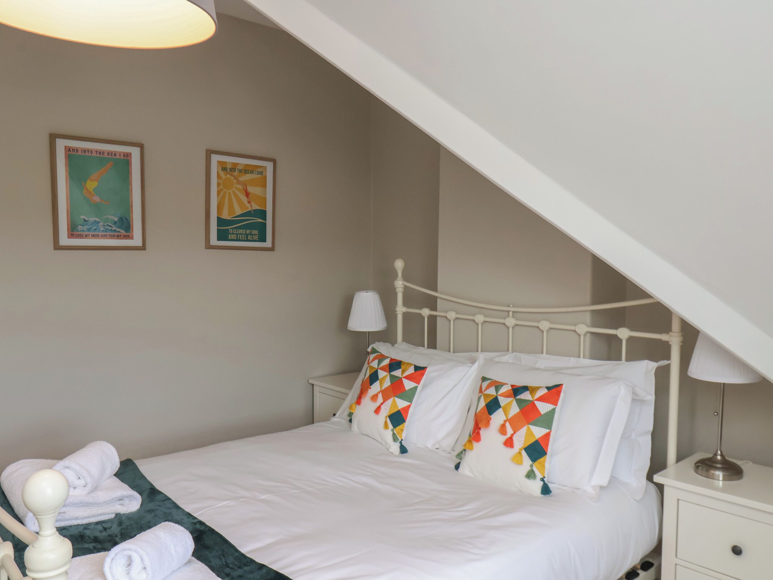 Un dormitorio con una cama blanca, almohadas coloridas, una mesita de noche blanca, una lámpara y dos carteles enmarcados en la pared.