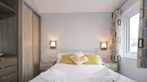 Una cama bien hecha con sábanas blancas y dos almohadas, un armario de madera, una mesita de noche con lámpara, y una ventana con cortinas.