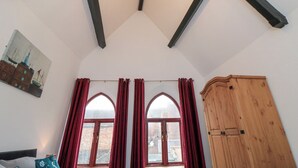 Une pièce avec un plafond en pente, des poutres en bois, un lustre, une armoire en bois et une fenêtre ornée de rideaux rouges.