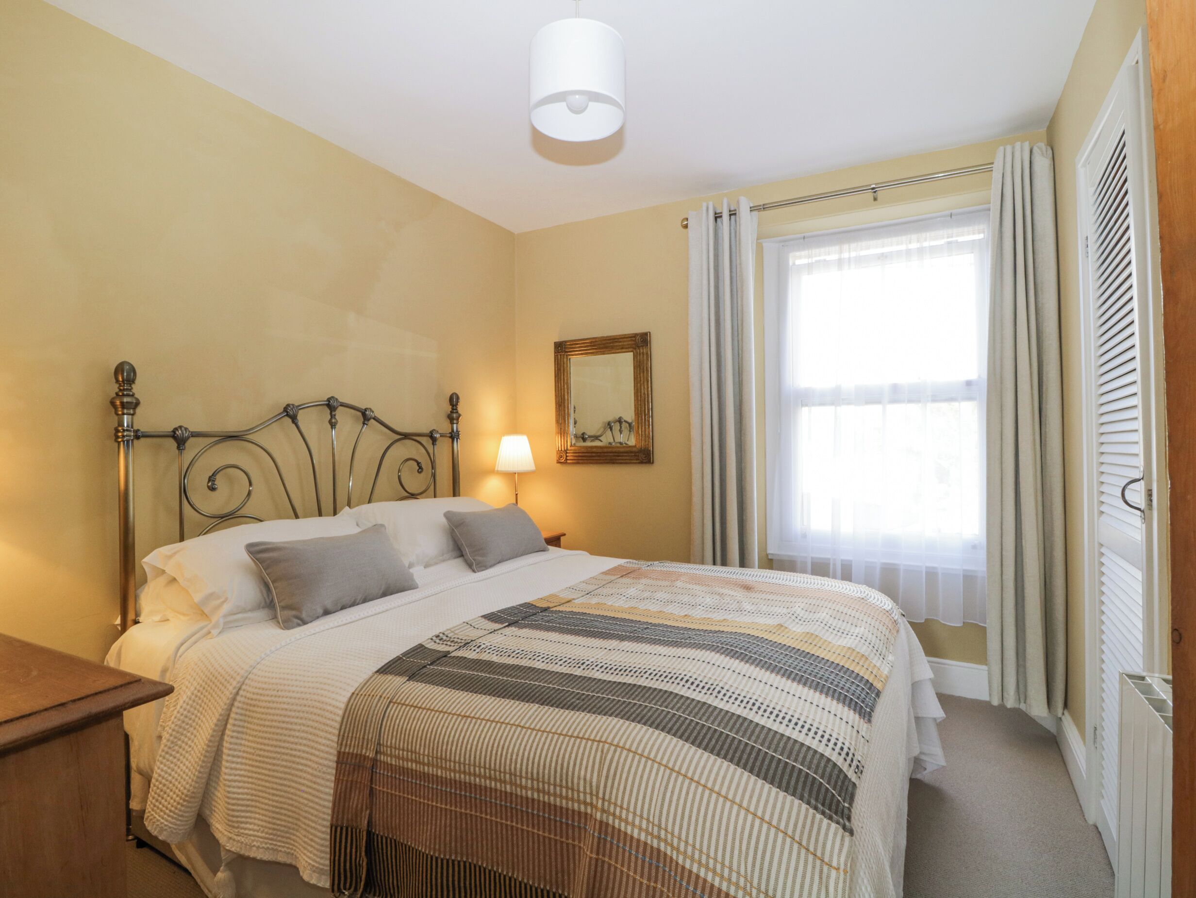 A bedroom with a bed, a bedside table, a lamp, a window with curtains, and a wall-mounted mirror.