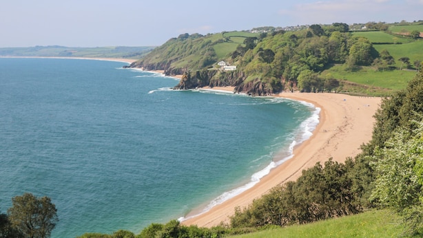 Un paysage côtier avec une plage de sable, des collines verdoyantes et une mer bleue et limpide.