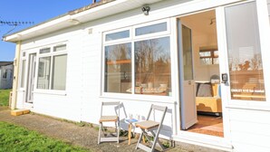Un bâtiment blanc d’un seul étage, doté d’une porte-fenêtre coulissante, d’un petit espace extérieur avec deux chaises et d’une fenêtre visible avec des stores.