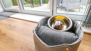 Ein Haustierbett mit einer Metallschüssel, beschriftet mit "Dog Treats", auf einem Holzboden neben einer Glastür.