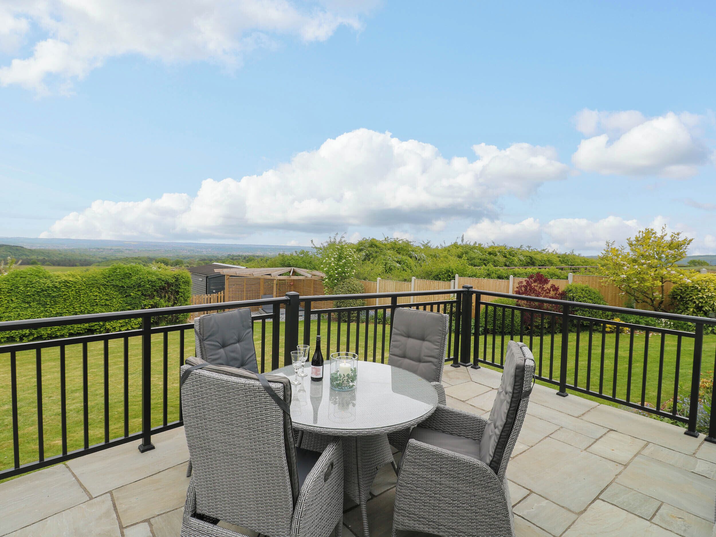 A patio with wicker furniture and a glass table, overlooking a green landscape and a distant view.