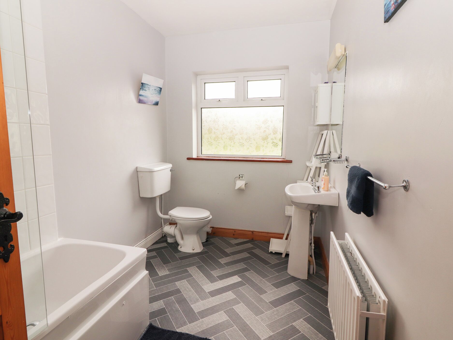 A small, clean bathroom with a toilet, sink, and bathtub. There is a window providing natural light and a radiator on the wall.