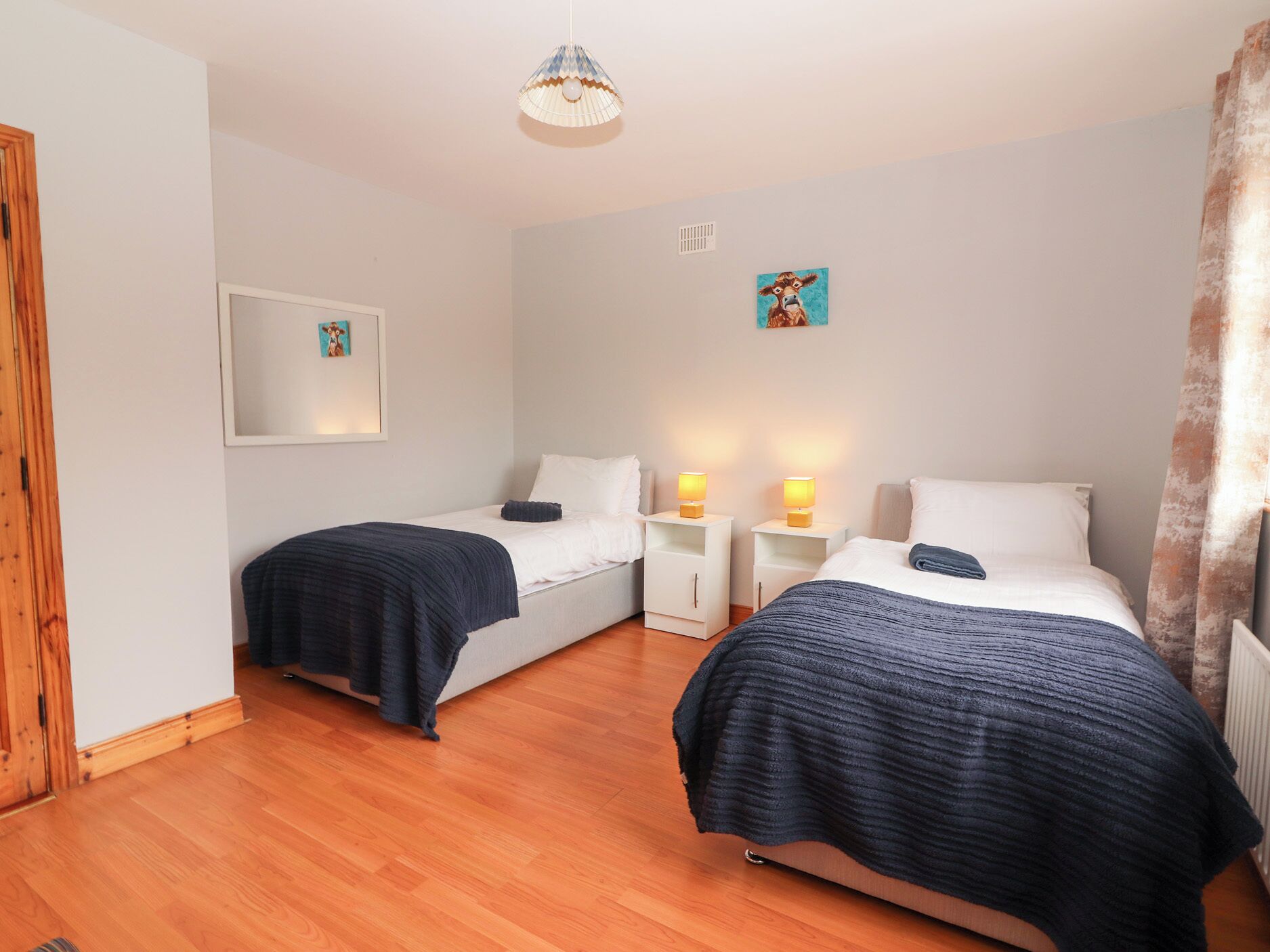 Una stanza con due letti, ognuno con una coperta blu scuro, cuscini bianchi e comodini con lampade. Si nota un quadro incorniciato appeso alla parete e una porta in legno sulla sinistra.