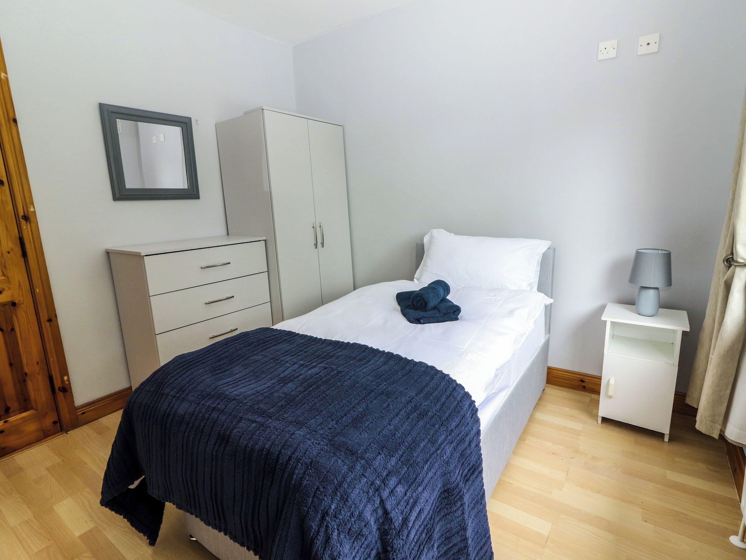 A bedroom with a bed, a wardrobe, a bedside table with a lamp, and a mirror on the wall.