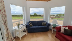 A room with a blue sofa, a red sofa, and a white table with a lamp. There are large windows with a view of a green landscape and mountains. - Cois Leice (Mulranny)