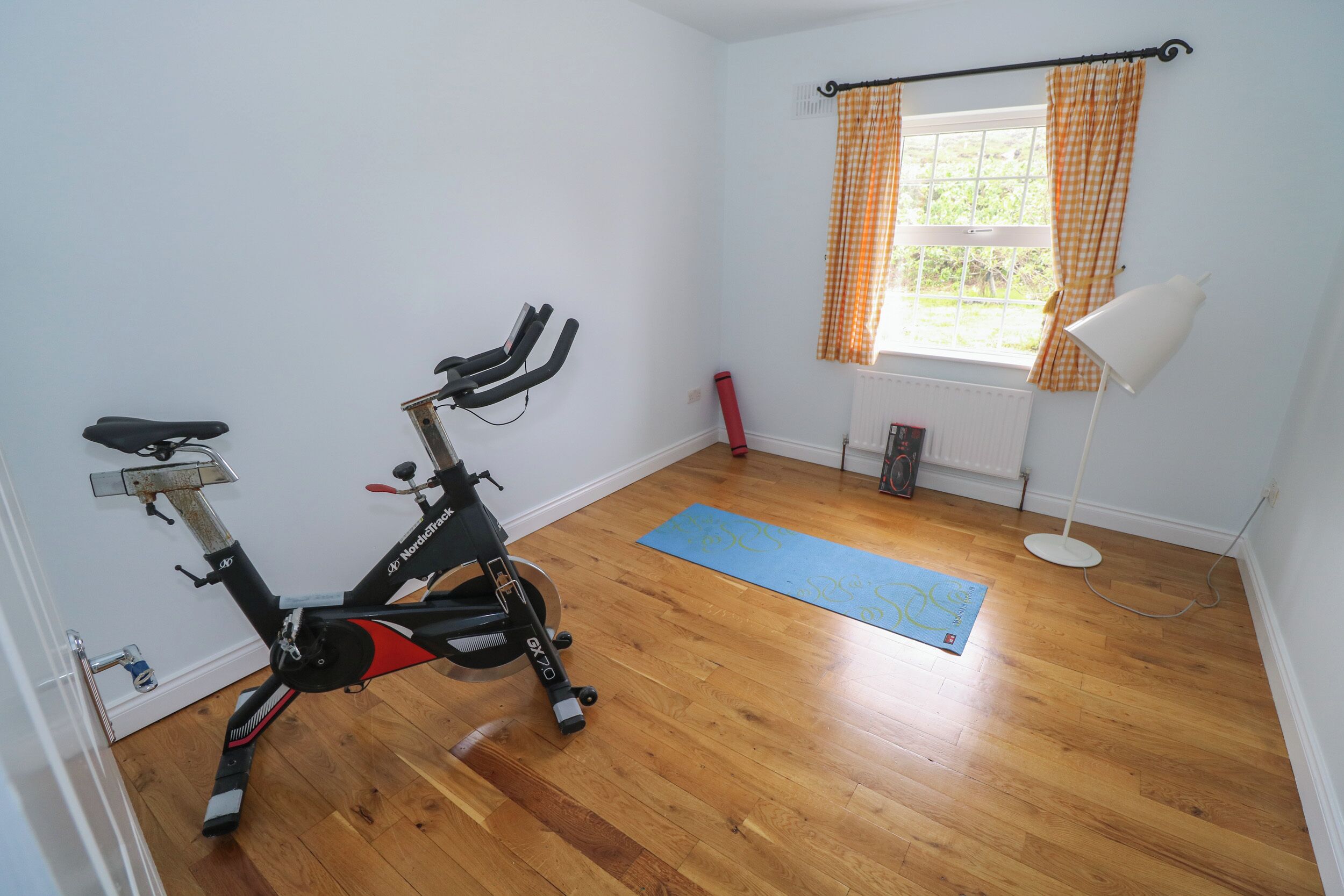 Una habitación con bicicleta estática, colchoneta de yoga, ventana con cortinas a cuadros y lámpara de pie.