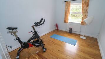 Una habitación con bicicleta estática, colchoneta de yoga, ventana con cortinas a cuadros y lámpara de pie.