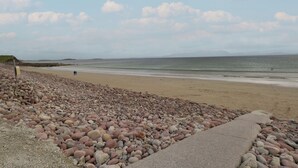 A pebbled beach with a concrete walkway, a distant view of the sea, and a small hill in the background. - Cois Leice (Mulranny)