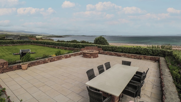 A patio with a table and chairs, a stone fire pit, and a view of the sea. - Cois Leice (Mulranny)