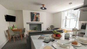 Ein modernes Wohnzimmer mit Essbereich, Kamin und einer durchgehenden Tür erkennbaren Küche.