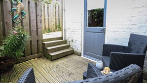 Eine Holzterrasse mit Korbmöbeln, einer Tür und einem kleinen Gartenbereich mit Stufen zu einem Tor.