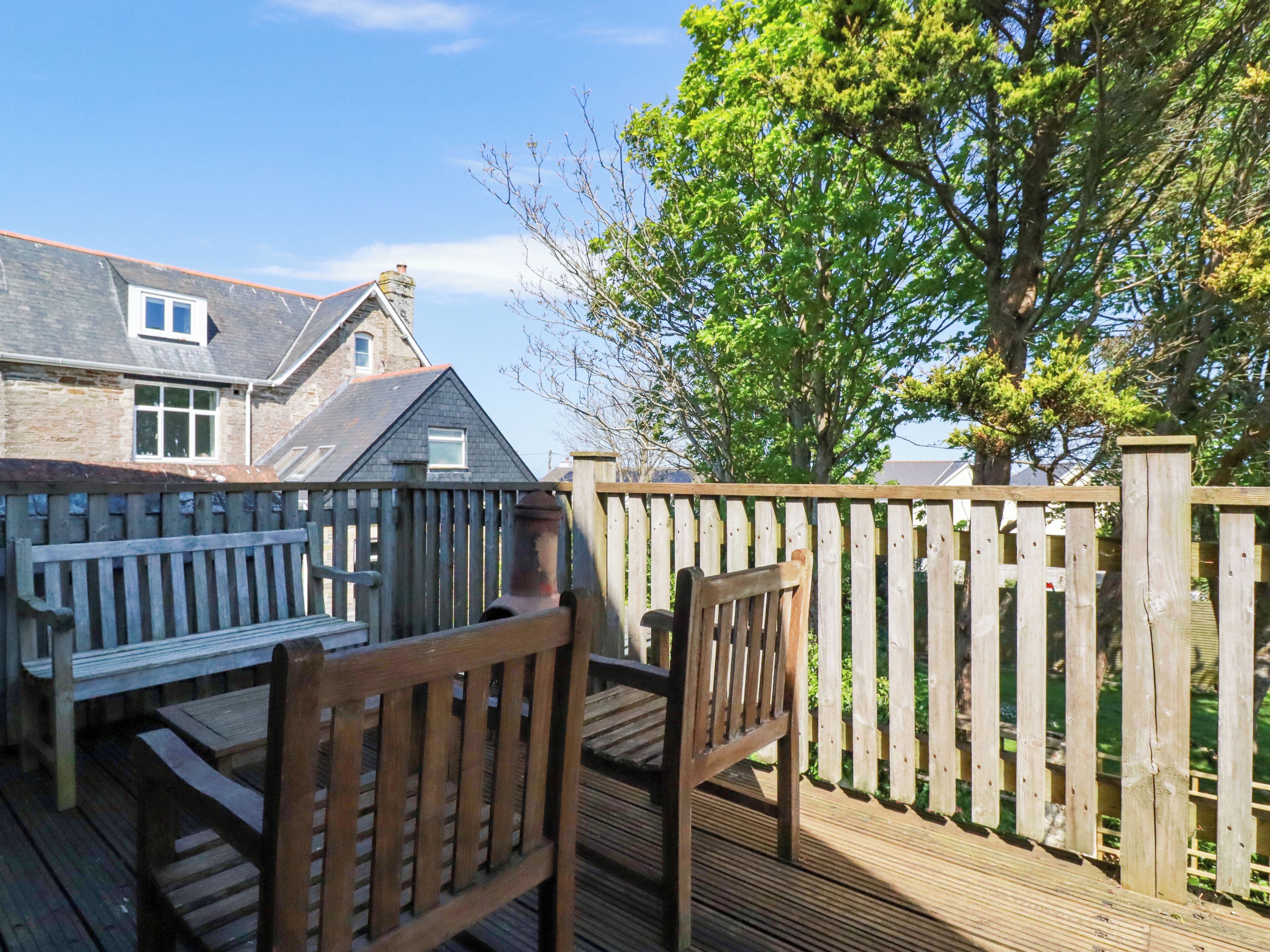 A wooden deck with a bench and chairs, a stone building with a chimney, and a tree in the background.